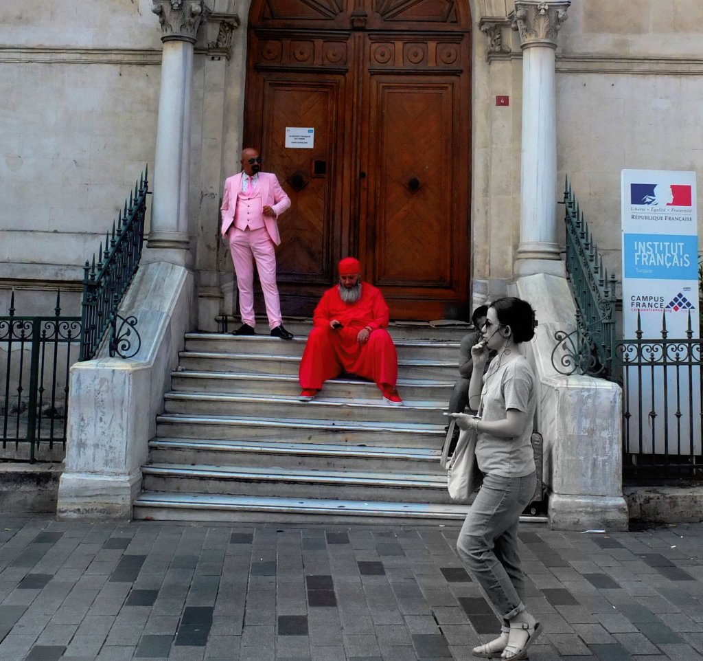 Entrance to the French Institute of Istanbul. A man is standing before the doorway, dressed smartly in a pink suit and tie. Another, bearded, man is seated, in what appears to be religious garb; his clothes and cap are bright red. Another man, also seated, is partly hidden by a woman passing by on the street. The photo has been retouched so that she will appear in monochrome.